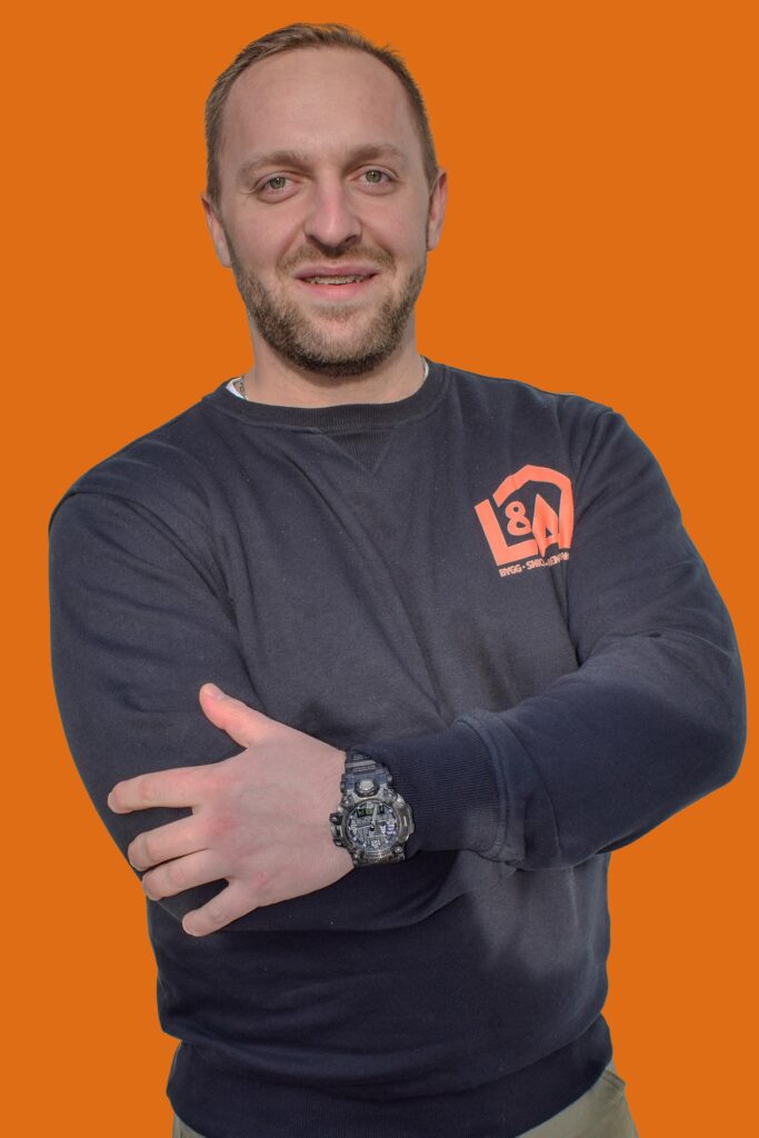 Artur Szynszecki, en av ägarna till L&A Fantastic Renovering i Stockholm, står med armarna i kors och ler mot kameran. Han bär en svart tröja med företagets logotyp och en robust armbandsklocka. Bakgrunden är orange.