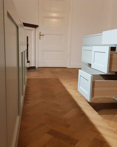 Pågående köksrenovering i Stockholm med ljusgrå skåp och öppna trälådor som visar hantverksmässig inredning. Köket har klassiska spegelluckor och står på ett vackert fiskbensmönstrat trägolv. En vit vacker dörr med mässingshandtag syns i bakgrunden.