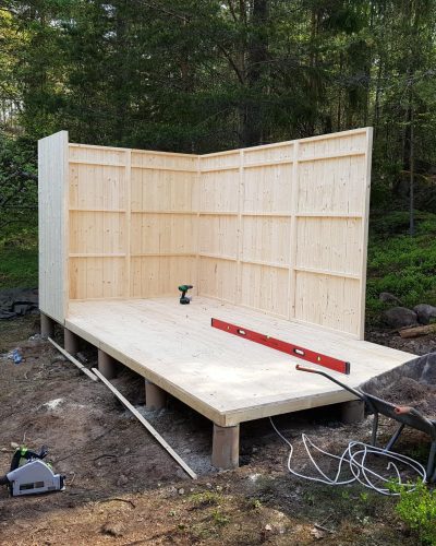 Byggnation av en träkonstruktion i skogen i Stockholms skärgård. En halvfärdig träbyggnad med golv och två väggar, omgiven av grön skog. Verktyg och material ligger utspridda runt bygget.