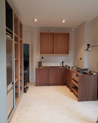 Pågående köksrenovering i Stockholm med mörka träunderskåp, öppna överskåp i ljust trä och integrerade vitvaror. Verktyg och material är framme under monteringen, och detaljer som handtag och bänkskivor saknas fortfarande. Vägglampa och infällda takspotlights ger en modern belysning.