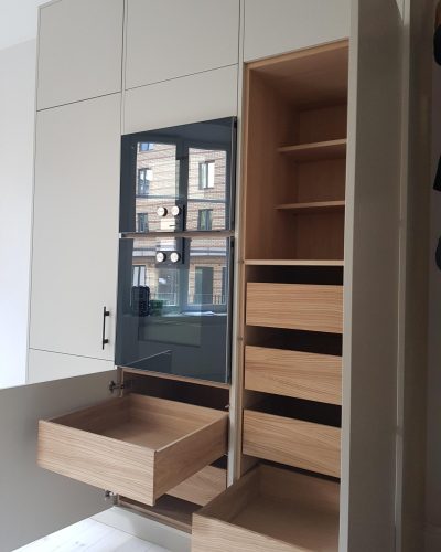 Nyrenoverat kök i Stockholm med ljusa skåp och integrerade svarta vitvaror. Öppna lådor och hyllor i ljust trä visar smart förvaring med en kombination av utdragslådor och öppna hyllplan. Spegling i ugnsluckan avslöjar en vy av byggnader utanför fönstret.