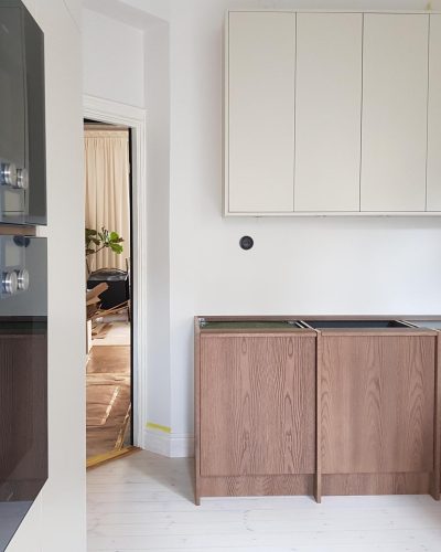 Pågående köksrenovering i Stockholm med ljusa överskåp, mörka träunderskåp och integrerade svarta vitvaror. Bänkskivor saknas fortfarande, och detaljer i rummet vittnar om pågående arbete. En öppning leder till ett vardagsrum med naturligt ljus och inredning.