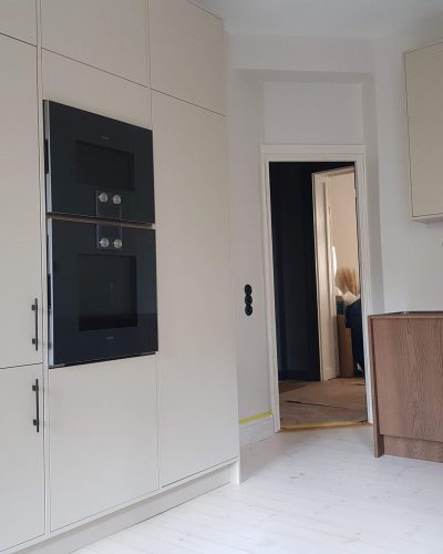 Stilrent renoverat kök i Stockholm med ljusa, släta skåpluckor och integrerade svarta vitvaror. Köket kombinerar moderna material med trädetaljer för en varm känsla. Svarta strömbrytare och handtag skapar en elegant kontrast mot de ljusa ytorna.