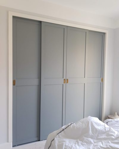 Platsbyggd garderob med grå skjutdörrar och mässingshandtag i ett ljust sovrum. Vita väggar och ett bäddat sängöverkast med svarta detaljer syns i förgrunden. Finsnickeri och renovering i Stockholm.