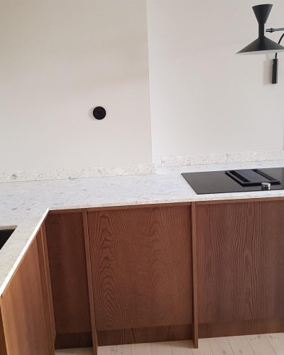 Nyrenoverat kök i Stockholm med mörka träskåp, ljus terrazzo-bänkskiva och svart inbyggd häll med integrerad fläkt. En svart vägghängd lampa ger modern belysning, och en svart diskho med mörk blandare kompletterar den stilrena designen.
