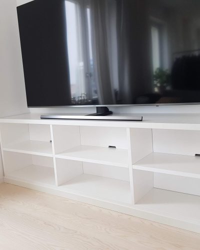 Platsbyggd vit TV-bänk med öppna hyllor och integrerade lådor, placerad i ett ljust rum med trägolv. En stor platt-TV står ovanpå bänken, och ett fönster med gardiner speglas i skärmen. Finsnickeri och renovering i Stockholm.