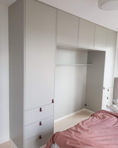 Platsbyggd garderob i ljusgrått med integrerad förvaring runt en sängplats. Garderoben har både skåp och lådor med stilrena läderhandtag. Ljust trägolv och en bäddad säng med rosa överkast syns i rummet. Finsnickeri och renovering i Stockholm.