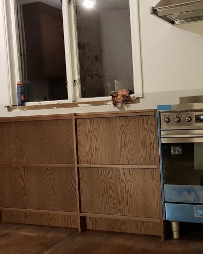 Pågående köksrenovering i Stockholm med mörka träskåp och en stor rostfri spis med skyddsfilm kvar. En vägghylla och fläkt är monterade, medan elinstallationer och fönsterdetaljer ännu är under arbete. Golvet är i mörkt trä för en varm känsla.