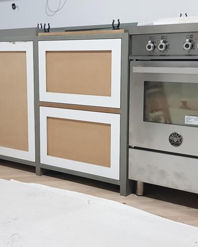 Pågående köksrenovering i Stockholm med halvfärdiga skåpluckor och en stor rostfri spis med dubbla ugnar. Köksinredningen har en klassisk design med inramade fronter, och skyddsplast täcker delar av golvet under arbetet.