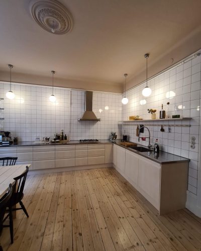 Renoverat kök i Stockholm med klassisk och modern design. Ljusa träluckor, rostfri bänkskiva och vit kaklad vägg ger en stilren känsla. Kökets öppna hyllor pryds av detaljer, medan takrosett och hängande lampor skapar en elegant atmosfär.