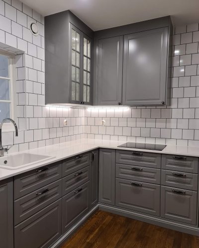 Nyrenoverat kök i Stockholm med gråa klassiska skåpluckor, vitrinskåp och vit kaklad vägg med mörk fog. Köket har en vit bänkskiva, porslinsho och moderna spotlights för belysning. Mörkt trägolv skapar en varm kontrast till de ljusa detaljerna.