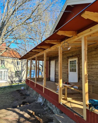 Renovering av en gammal veranda i Stockholm. En pågående renovering av en veranda med ny träkonstruktion. Den gamla timmerväggen är synlig, och omgivningen består av ett äldre hus och träd.