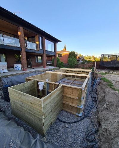 Pågående konstruktion av en pool och trädäck i Stockholm. En trästruktur byggs som en del av ett poolområde utanför en modern villa. Jorden runt konstruktionen är grävd, och rördragningar syns vid sidan av.