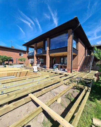 Trädäck byggs vid en modern villa i Stockholm. En större trästomme för ett nytt trädäck håller på att resas framför en tegelvilla med stora fönster. Två hantverkare arbetar i solen.