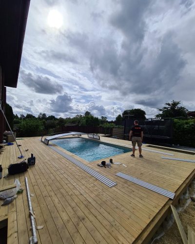 Pooldäck under konstruktion i Stockholm – Ett stort trädäck byggs runt en pool, med en hantverkare som arbetar på plats. Olika verktyg och byggmaterial ligger utspridda på trädäcket under en molnig himmel.