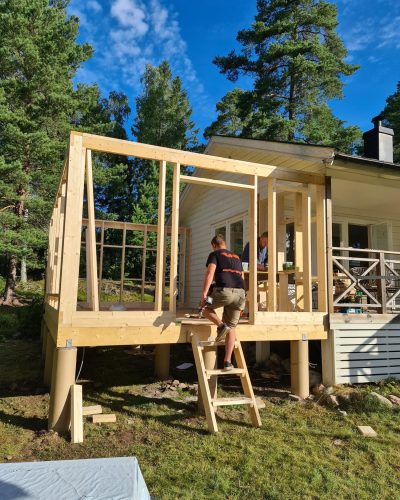 Pågående tillbyggnad av ett hus i Stockholm. En hantverkare klättrar upp för en provisorisk trästege till den nya delen, som är uppförd på plintar och har en synlig trästomme. En annan hantverkare arbetar inuti konstruktionen. Huvudbyggnaden är vit med en altan och omgivningen är grön med höga träd och blå himmel.