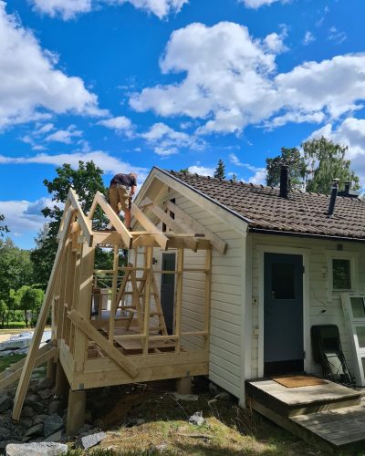 Tillbyggnad under konstruktion på ett hus i Stockholm. En hantverkare arbetar på takstommen till den nya delen, som är byggd på plintar och har en synlig trästomme. Huvudbyggnaden är vit med en blå dörr och ett tegeltak. Omgivningen består av grönska, träd och en klarblå himmel med vita moln.