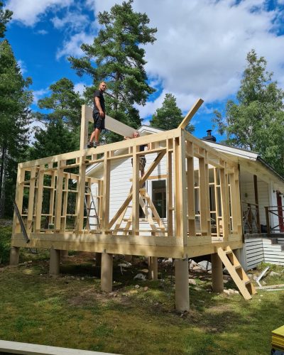 Pågående byggnation av en tillbyggnad på ett hus i Stockholm. Två hantverkare arbetar med trästommen, varav en står på takbjälkarna. Tillbyggnaden är upphöjd på plintar och har en synlig trästomme. Huvudbyggnaden är vit med en veranda och röda dörrar. Omgivningen består av grönska och höga träd, med en blå himmel och vita moln i bakgrunden.