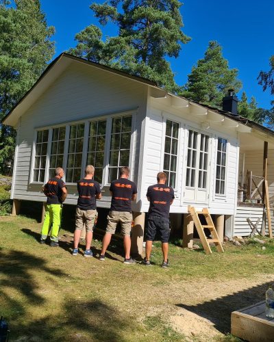Fyra snickare i svarta tröjor från L&A Fantastic Renovering med företagslogga står framför en nybyggd vit tillbyggnad på ett hus i Stockholm. Tillbyggnaden har stora fönster och är upphöjd på plintar. Runt huset finns byggmaterial och verktyg, och en trätrappa leder upp till verandan. Omgivningen består av grönskande träd och en klarblå himmel.