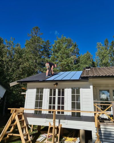 Takrenovering på ett vitt trähus i Stockholm. En hantverkare arbetar på taket och monterar ny takbeläggning bredvid befintliga takpannor. Huset har stora fönster och en tillbyggnad under konstruktion, med byggställningar och trästöd runtom. Bakgrunden består av grönskande träd och en klarblå himmel.