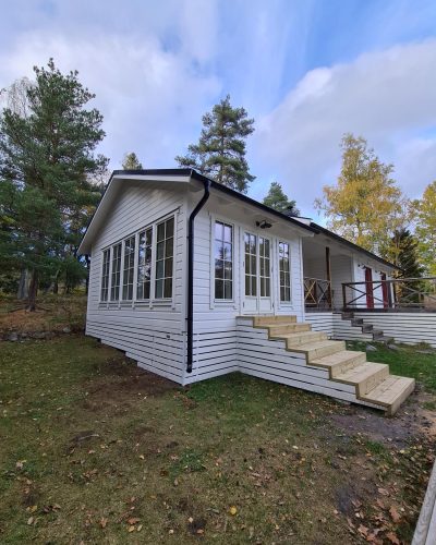 Nyrenoverad vit stuga i Stockholm med stora fönster och en ny trappa i trä som leder upp till ingången. Omgiven av grönska med tallar och lövträd i bakgrunden. Huset är en utbyggnad av en befintlig bostad med en veranda och räcke i trä.