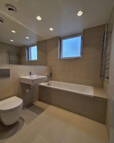 Modern badrumsrenovering i Stockholm med beige kakel på väggar och golv. Badrummet har ett vägghängt handfat, en inbyggd badkar med dusch, samt en vägghängd toalett. Två frostade fönster släpper in naturligt ljus, och en stor spegel täcker en del av väggen. Taket är utrustat med infällda spotlights för jämn belysning.