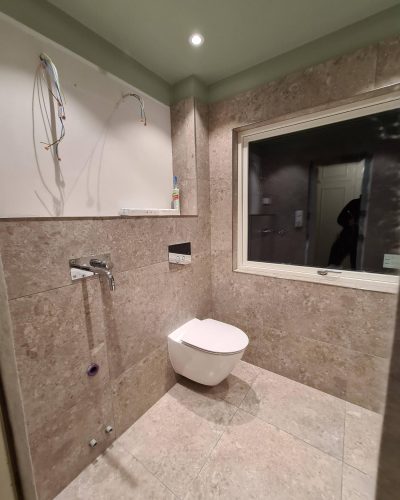 Pågående badrumsrenovering i Stockholm med vägghängd toalett. Väggar och golv är klädda i beige naturstensliknande kakel, och taket är målat i en mjuk grön nyans. Infällda spotlights ger belysning. Ovanför toaletten syns en vägg med dragna elledningar, förberedda för installation av en spegel eller badrumsskåp.