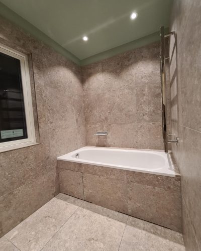 Badrumsrenovering i Stockholm med inbyggt badkar. Väggar och golv är klädda i naturstensliknande kakel i beige nyans, medan taket har en mjuk grön färg. Infällda spotlights ger stämningsfull belysning, och en vägghängd handdukstork i krom finns vid sidan av badkaret.