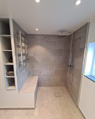Badrumsrenovering i Stockholm med rymlig duschhörna. Väggar och golv är klädda i beige naturstensliknande kakel. En inbyggd sittbänk och vägghängd handdukstork finns på ena sidan, medan en stor takdusch är monterad på den motsatta väggen. Inbyggda hyllor ger extra förvaringsutrymme.