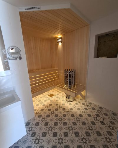 Renovering av badrum med inbyggd bastu i Stockholm. Bastun är klädd i ljust trä med en modern bastuugn och väggbelysning. Badrummet har ett mönstrat klinkergolv i grå och vita nyanser samt vita kaklade väggar. Ett vitt tvättställ syns till vänster, och en inbyggd nisch i väggen ger extra förvaringsutrymme.