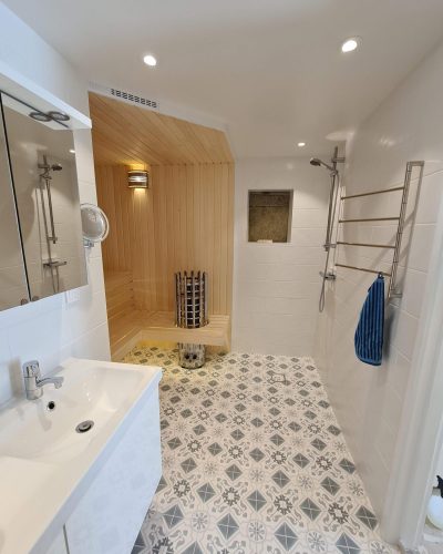 Badrumsrenovering i Stockholm med inbyggd bastu. Badrummet har mönstrat klinkergolv i grå och vita nyanser, vita kaklade väggar och en dusch med glasvägg. Bastun är klädd i ljust trä med en modern bastuugn. Ett vitt tvättställ med spegelskåp och en kromad handdukstork med blå handduk kompletterar det stilrena uttrycket.