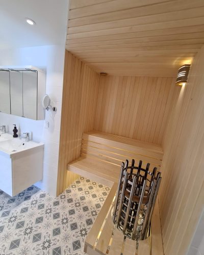 Nyrenoverat badrum med integrerad bastu i ljust trä. Bastun har bänkbelysning och en modern, rostfri bastuugn. Badrummet har mönstrat klinkergolv, vita kaklade väggar och en stilren tvättställskommod med spegelskåp. Badrumsrenovering i Stockholm.