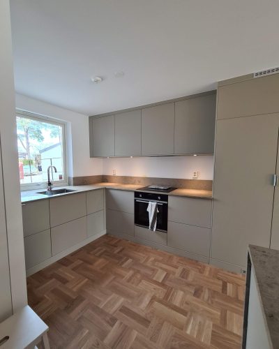 Stilrent renoverat kök i Stockholm med ljusgrå släta skåpluckor, stenskiva i naturton och integrerad ugn. Fiskbensmönstrat parkettgolv ger en varm kontrast till de moderna ytorna. Stort fönster vid diskhon släpper in naturligt ljus.