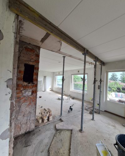 Pågående renovering av en bostad i Stockholm, där en vägg har rivits och en öppning förstärkts med en träbalk. Stödproppar håller upp taket under arbetet. Exponerade tegelstenar och byggmaterial ligger på golvet. Stora fönster i bakgrunden ger utsikt mot en grönskande trädgård med utemöbler.