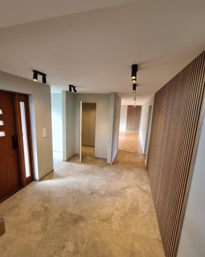 Nyrenoverad hall i en bostad i Stockholm med ljusa väggar, stora beige klinkerplattor på golvet och en träbeklädd vägg med vertikala ribbor. Svarta takspotlights ger belysning, och en brun träytterdörr syns till vänster. Hallen leder vidare till rum med parkettgolv.