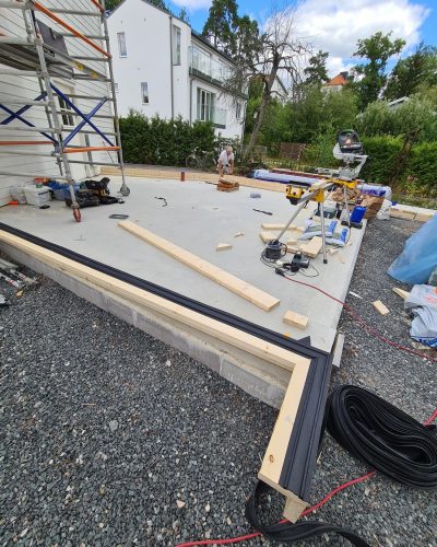 Byggarbetsplats i Stockholm där en betongplatta för en ny byggnad har gjutits. Träreglar och fuktskydd monteras längs kanten. Byggställningar står intill en befintlig vit träfasad. En hantverkare arbetar på plattan, och verktyg som kap- och gersåg samt elutrustning är utspridda. I bakgrunden syns moderna villor och grönska under en delvis molnig himmel.