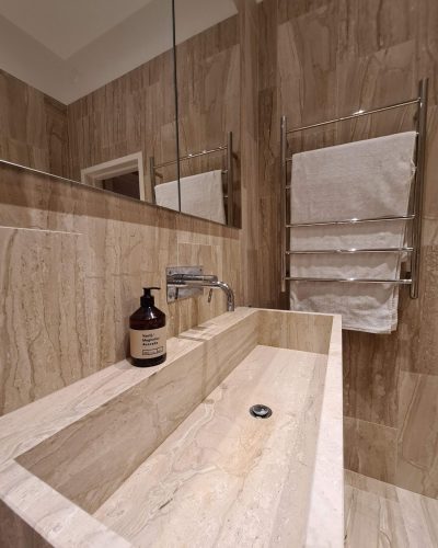 Stilren badrumsrenovering med marmorinspirerat handfat i ett stycke. Väggarna är klädda i beige naturstensliknande kakel, och ovanför tvättstället sitter en stor spegel. En kromad handdukstork med en vit handduk bidrar till den eleganta känslan.