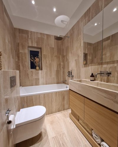 Elegant badrumsrenovering med naturinspirerad design. Väggar och golv är klädda i beige marmorliknande kakel, och trädetaljer i kommoden skapar en varm känsla. Det vägghängda tvättstället har en stilren marmorskiva, och badkaret är integrerat med en regndusch ovanför. Ett litet fönster med dekorativa växter släpper in naturligt ljus.