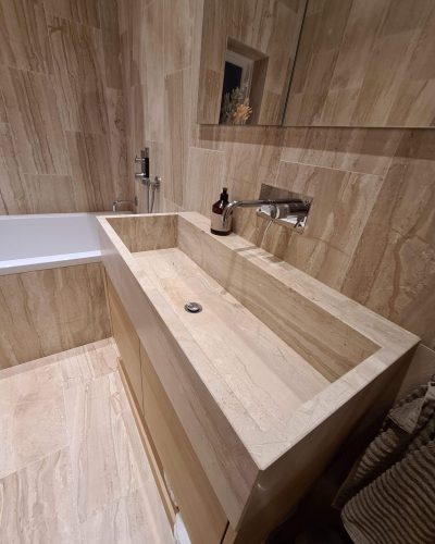Lyxigt badrum i naturtoner med väggar, golv och handfat i beige marmor. Det stilrena handfatet har en minimalistisk design med vägghängd kran i krom. Intill finns ett badkar med inbyggd dusch. En spegel ovanför handfatet reflekterar rummets eleganta detaljer. Badrumsrenovering med exklusiva materialval.
