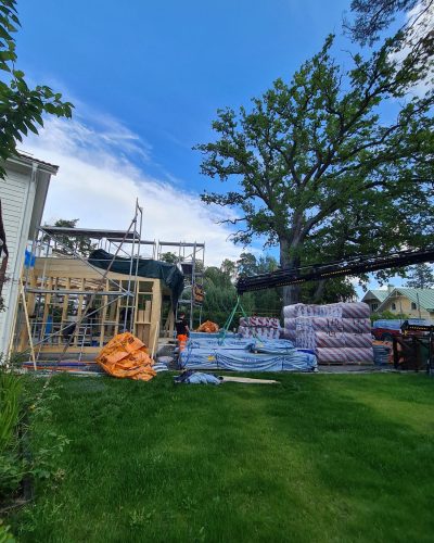 Pågående tillbyggnad i Stockholm med trästomme under uppförande. Byggställningar omger konstruktionen, och byggmaterial som isolering och virke är staplat på gräsmattan. En kranlastbil lyfter material, och byggarbetare i varselkläder syns på plats. En stor ek reser sig i bakgrunden, och himlen är blå med några moln.