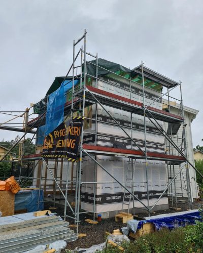 Pågående byggprojekt i Stockholm där en husfasad är under renovering och utbyggnad. Ställningar omger byggnaden, och väggarna är täckta med byggfolie och ångbroms. En skylt med "Byggsäker Renovering" syns på ställningen. Byggmaterial finns utplacerade på arbetsplatsen.