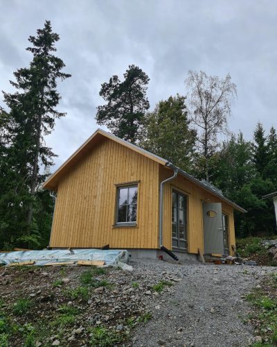 Nybyggt trähus i Stockholm under totalentreprenad med fasad i ljus träpanel och ett gråmålat dörrparti. Huset står på en grusad tomt omgiven av skog och grönska. Byggmaterial och plastskydd ligger på marken, vilket tyder på att byggprojektet ännu inte är helt färdigställt.