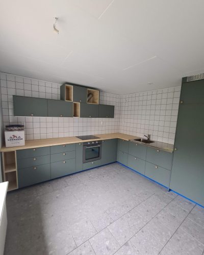 Nyrenoverat kök i Stockholm med grönmålade skåp, ljusa trädetaljer och vitkaklad vägg. Köket har en integrerad ugn, en stilren diskho och bänkskiva i trä. Golvet är lagt med grå kalkstensplattor för en modern och tidlös känsla.