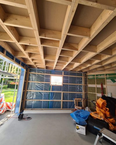 Pågående byggnation av en träkonstruktion i Stockholm. Bilden visar ett garage under uppförande med synlig träbjälklagstak, plastfolie och isolering på väggarna. Byggmaterial, verktyg och presenningar ligger på betonggolvet. Öppningen leder ut till en grönskande trädgård.