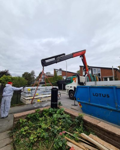 En lastbil med kran lyfter byggmaterial från Beijer Byggmaterial vid en renovering i Stockholm. En arbetare i vit skyddsoverall och röd keps guidar lasten. I förgrunden syns en blå container märkt "LOTUS" och en rabatt med grönska. I bakgrunden finns radhus med röda tegelväggar och svarta tak.