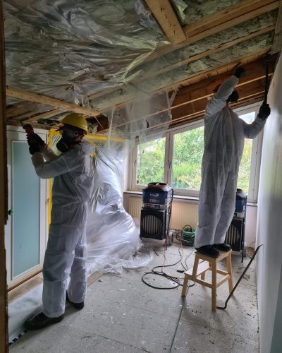 Två hantverkare i skyddsutrustning, inklusive vita overaller, hjälmar och andningsmasker, arbetar med att riva taket i ett rum under renovering i Stockholm. Isolering och plast hänger från takbjälkarna, och byggdamm täcker golvet. Två industriella luftrenare står vid fönstret, där gröna träd syns utanför.