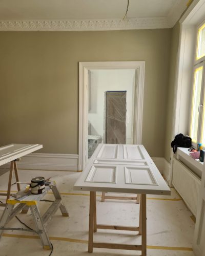 Pågående renovering i ett sekelskiftesrum med målade väggar i beige ton och dekorativa taklister. Två spegeldörrar ligger på bockar för målning, och golvet är täckt med skyddspapper. Renovering i Stockholm.