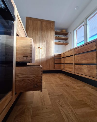 Exklusiv köksrenovering i Stockholm med skräddarsydda trälådor i ljus ek och detaljerad sinkfog. Köket har integrerade vitvaror, öppna hyllor och ett stort skafferi i samma träslag. Fiskbensmönstrat parkettgolv och stora fönster ger en ljus och varm atmosfär.