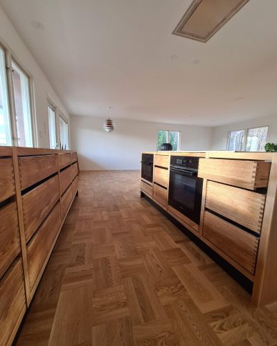 Nyrenoverat kök i Stockholm med köksöar i ljus ek och integrerade ugnar. Köket har stilrena lådor med sinkfog, parkettgolv i fiskbensmönster och stora fönster som släpper in naturligt ljus. Öppen planlösning mot vardagsrum.