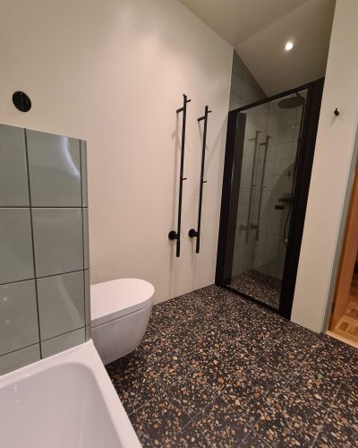 Stilrent renoverat badrum med terrazzo-golv i mörka och beige toner. Väggarna är delvis kaklade med gröna plattor och en modern vägghängd toalett sitter mot en ljusmålad vägg. Svarta detaljer som handdukshängare och duschväggens ram förstärker den minimalistiska designen. Glasdörren leder in till en dusch med regnduschmunstycke. Badrumsrenovering i Stockholm.